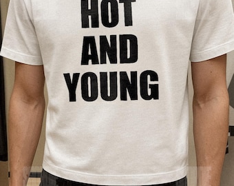 Könnte beinhalten: Weißes T-Shirt mit dem Aufdruck "HOT AND YOUNG" in großen, schwarzen Buchstaben. Das Shirt hat kurze Ärmel und einen Rundhalsausschnitt. Der Text ist mittig auf der Vorderseite platziert.