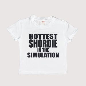 Puede incluir: Camiseta blanca con texto negro que dice "HOTTEST SHORDIE IN THE SIMULATION"