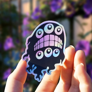 May include: A die-cut sticker featuring a dark, amorphous creature with multiple eyes and a wide, toothy grin. The design incorporates a holographic finish that reflects light, creating a shimmering, iridescent effect across the surface.