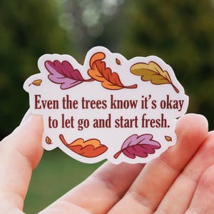 May include: A white die-cut sticker featuring the dark red text Even the trees know it's okay to let go and start fresh. The design is decorated with colorful autumn leaves in shades of orange, yellow, and purple surrounding the lettering.