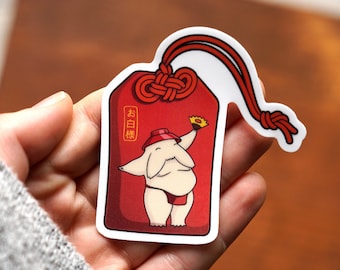 May include: Red and white Japanese charm with a cartoon character wearing a red hat and holding a flower. The charm has the text "お白様" written on it.