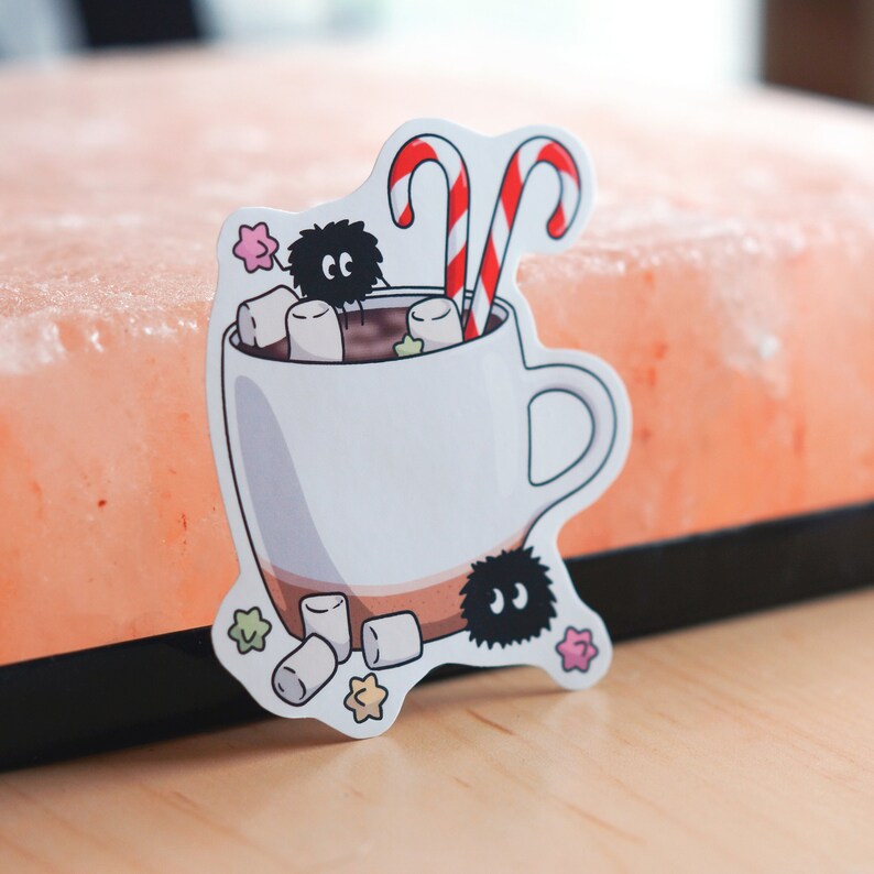 May include: A white mug with a brown rim filled with hot chocolate, marshmallows, and candy canes. Two black and white fuzzy creatures are peeking out of the mug. The mug is on a pink and white background.