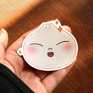 Puede incluir: Un dumpling de dibujos animados blanco con una cara feliz y los ojos cerrados. El dumpling tiene un rubor en las mejillas y sonríe con la boca abierta. Hay dos pequeñas estrellas amarillas sobre la cabeza del dumpling.