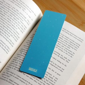 May include: A blue bookmark with a gradient design and small white dots, placed in a book. The bookmark has the text "NATALIE B DESIGNS" at the bottom.