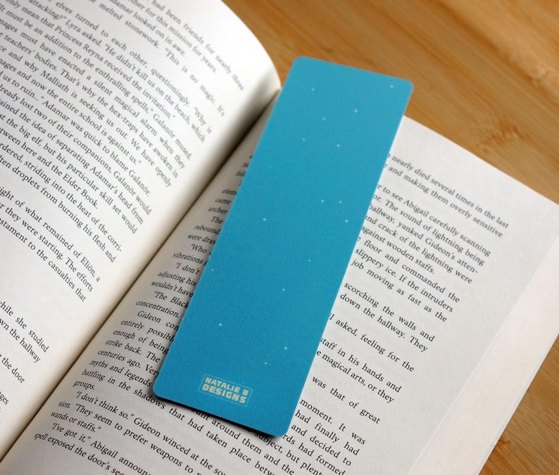 May include: A blue bookmark with a gradient design and small white dots, placed in a book. The bookmark has the text "NATALIE B DESIGNS" at the bottom.