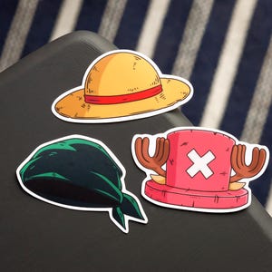 May include: Three cartoon stickers featuring items from the anime series One Piece. The stickers include a yellow straw hat with a red band, a dark green bandana, and a pink hat with antlers and a white cross.