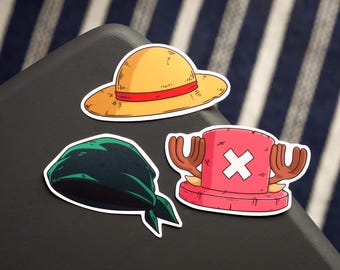 May include: Three cartoon stickers featuring items from the anime series One Piece. The stickers include a yellow straw hat with a red band, a dark green bandana, and a pink hat with antlers and a white cross.