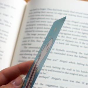 May include: A light blue bookmark featuring a cartoon ghost illustration and yellow star accents. The bookmark is held in front of an open book with printed text. The bookmark is approximately 15 cm long.