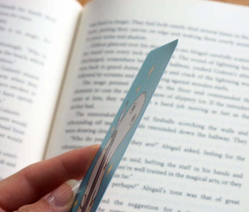 May include: A light blue bookmark featuring a cartoon ghost illustration and yellow star accents. The bookmark is held in front of an open book with printed text. The bookmark is approximately 15 cm long.