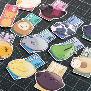 May include: A selection of vibrant stickers depicting cartoon animals, each accompanied by a rectangular tag. The tags feature Japanese text and are in different colours. The stickers are arranged on a black cutting mat.