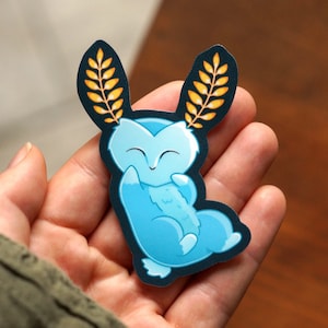 May include: A blue sticker featuring a cartoon fox-like creature with large ears adorned with yellow wheat. The creature is smiling and hugging itself. The sticker has a dark blue border.