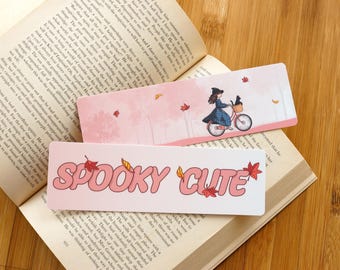 May include: Two rectangular bookmarks are displayed on an open book. One shows a girl on a bicycle with a cat. The other has the words "SPOOKY CUTE" in pink with autumn leaves.
