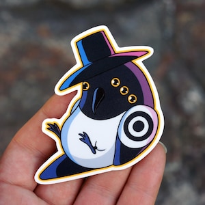 May include: A sticker featuring a cartoon bird wearing a top hat. The bird has a black body with white and blue accents, three yellow eyes, and a circular design on its side. The hat is black with blue and pink trim.