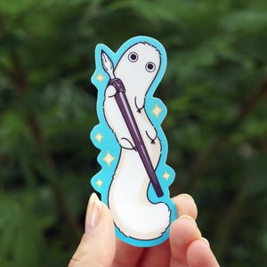 Op de afbeelding: Een sticker met een wit, cartoonachtig wezen met grote ogen, dat een penseel vasthoudt. De sticker heeft een lichtblauwe rand met gele steraccenten. Het wezen heeft een lange, pluizige staart.