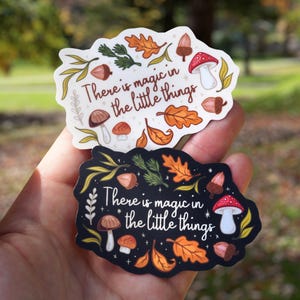 May include: Two whimsical stickers with the phrase "There is magic in the little things." One sticker is white with fall-themed illustrations, the other is black. Both feature mushrooms, acorns, and leaves.