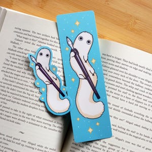 May include: Two whimsical bookmarks featuring a white creature holding a pen, set against a light blue background with gold stars. One bookmark is a die-cut shape, the other is rectangular. The bookmarks are placed on an open book.