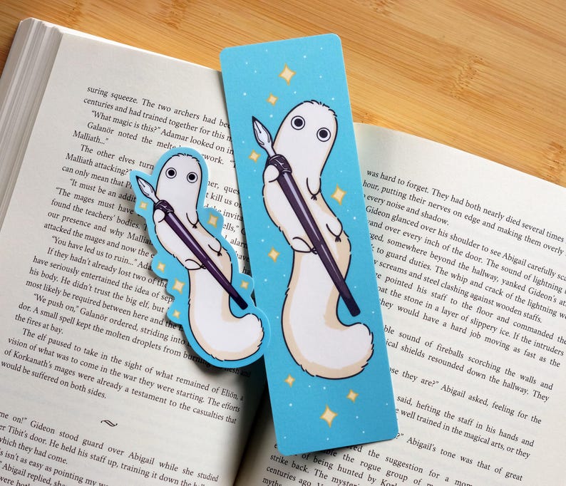 May include: Two whimsical bookmarks featuring a white creature holding a pen, set against a light blue background with gold stars. One bookmark is a die-cut shape, the other is rectangular. The bookmarks are placed on an open book.