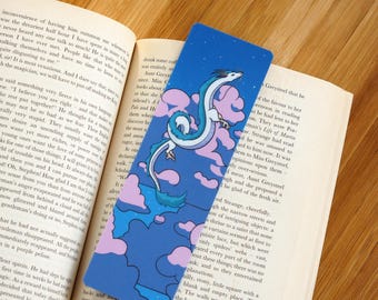 May include: A blue bookmark with a white and teal dragon design and pink and blue clouds. The bookmark is rectangular and is placed in an open book. The dragon design is on the front.