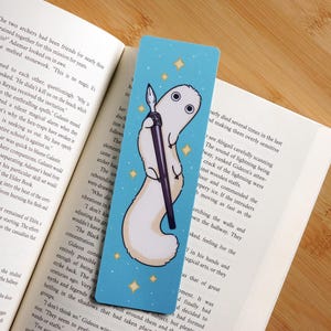May include: A blue bookmark featuring a white cartoon creature holding a black pen. The bookmark has a light blue background with yellow star accents. The bookmark is placed on top of an open book.