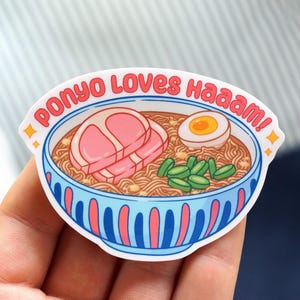 Pu&ograve; includere: Un adesivo raffigurante una ciotola di ramen con noodles, una fetta di maiale, un uovo sodo e verdure verdi. Il testo "Ponyo Loves haaaam!" &egrave; scritto in rosso intorno alla ciotola.