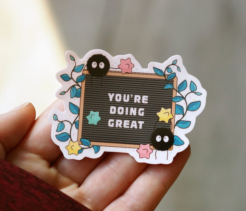 May include: A sticker with a black letter board that says "You're doing great". The letter board is surrounded by a vine with leaves and flowers. There are two black fuzzy creatures with big eyes on the sticker.