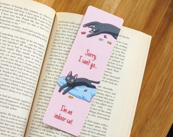 May include: A pink bookmark featuring a black cat illustration and the text "Sorry, I can't go..." and "I'm an indoor cat." The cat is on a blue cushion. The bookmark is placed in an open book.