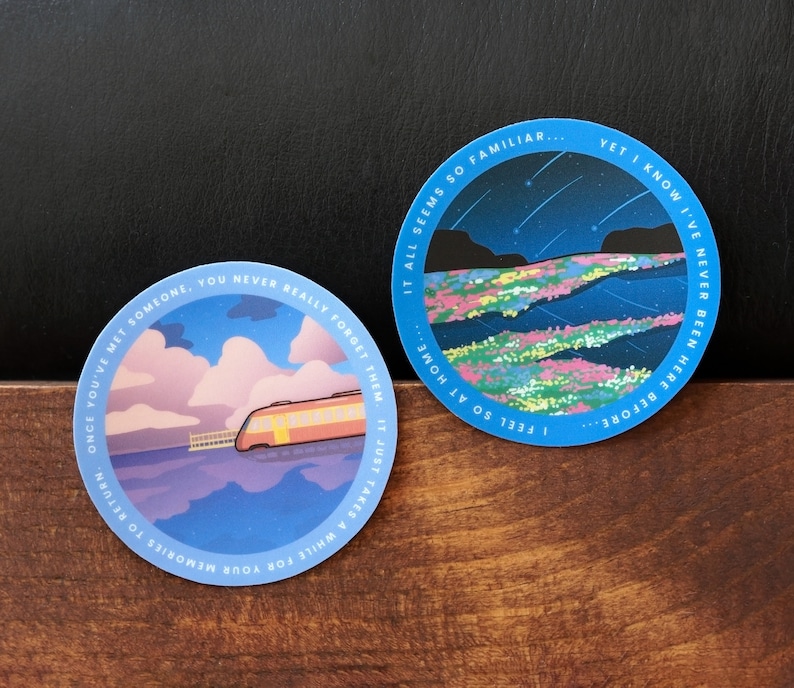 May include: Two blue circular stickers with white borders. The sticker on the left features a train traveling along a body of water with a pink and blue sky. The text on the sticker reads "Once you've met someone, you never really forget them. It just takes a while for your memories to return." The sticker on the right features a night sky with stars and a body of water with a colorful landscape. The text on the sticker reads "I feel so at home... It all seems so familiar... Yet I know I've never been here before."