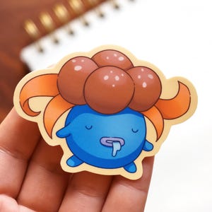 Peut inclure: Un autocollant découpé représentant un personnage de dessin animé avec un corps bleu, une tête bulbeuse marron et des détails orange. Le personnage a une bave et une expression neutre. L'autocollant est tenu dans une main.