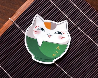 May include: A white cat with orange and gray markings is inside a green bowl. The cat is smiling and has its tongue sticking out. The bowl has Japanese characters on it.
