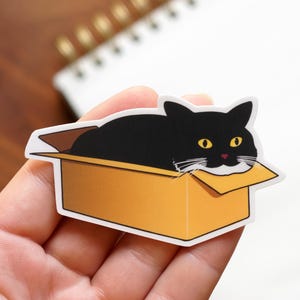 Puede incluir: Una pegatina que representa un gato negro con ojos amarillos y nariz rosa, asomando por una caja de cartón amarilla. El gato está dibujado en estilo de dibujos animados con bigotes blancos. La pegatina tiene un borde blanco.
