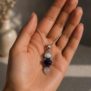 May include: A silver pendant necklace with three gemstones: a white moonstone, a purple amethyst, and a pink rose quartz. The pendant hangs from a fine silver chain. The gemstones are oval and teardrop shaped.