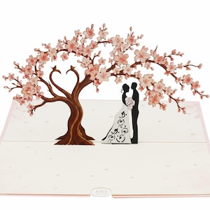 Könnte beinhalten: Eine Pop-up-Grußkarte mit einem dreidimensionalen Papierschnitt eines großen Kirschblütenbaums mit rosa Blüten und einem braunen Stamm. Unter den Ästen steht eine schwarze Silhouette von zwei Figuren, die sich gegenüberstehen.