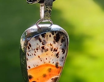 Op de afbeelding: Een Montana mosagaat hanger gezet in 925 sterling zilver, met een druppelvormige steen met oranje en witte banden en donkere insluitingen. De hanger hangt aan een decoratieve, getextureerde sterling zilveren hangerhouder en ketting.