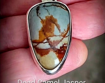 Op de afbeelding: Een druppelvormige Dead Camel Jaspis hanger gezet in een gepolijste zilveren zetting met een getextureerd zilveren oogje. De steen heeft een glad, meerkleurig oppervlak met wervelende patronen van beige, crème en dieproodbruine aders.