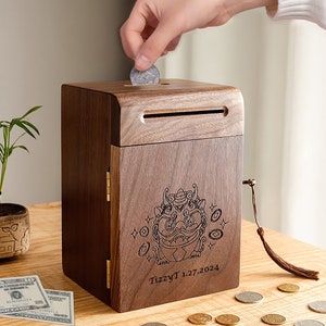 Peut inclure: Une tirelire en bois avec un motif de lion et le texte "TizzyT 1.27.2024" grav&eacute; sur le devant. La tirelire a une fente sur le dessus pour ins&eacute;rer des pi&egrave;ces de monnaie et un petit pompon suspendu sur le c&ocirc;t&eacute;.