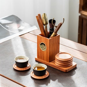 Könnte beinhalten: Ein hölzernes Teeset mit einem Ständer, einer Tasse, einer Untertasse und einem Halter für Teewerkzeuge. Der Halter hat ein dekoratives Wellenmuster. Das Set steht auf einem braunen Tisch.