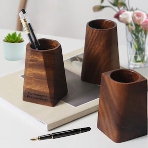May include: Three wooden pen holders, two are cylindrical and one is a geometric shape. The geometric pen holder has the words "Customized Personalized" engraved on the side.