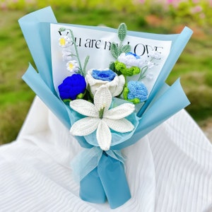 May include: A blue and white crocheted flower bouquet with a white lily, blue roses, and white baby's breath. The bouquet is wrapped in blue tissue paper and tied with a white ribbon. The bouquet has a card that says "You are my love".