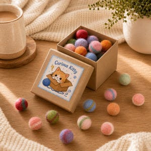 May include: A square cardboard box filled with small, multicoloured felt balls. The lid features a sticker with a cartoon orange cat in a box and the text Curious Kitty. Several additional felt balls in shades of pink, blue, green, and orange are scattered on the surface around the box.