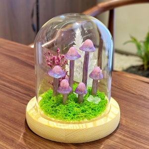 May include: A glass dome with a wooden base contains a miniature forest scene.  The scene features a bed of green moss and six purple mushrooms with white accents.