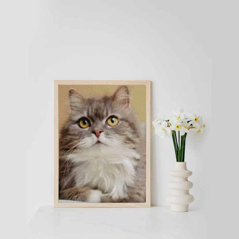 May include: A framed print of a grey and white cat with yellow eyes looking upwards. The cat is sitting in a relaxed pose with its paws tucked under its body. The frame is made of wood and has a light brown finish.