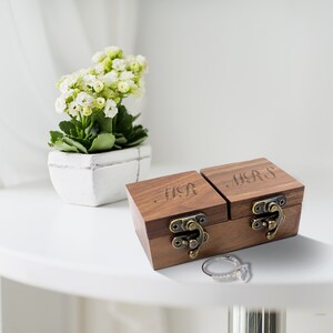 Op de afbeelding: Twee houten ringdozen met gegraveerde deksels. De doos aan de linkerkant zegt "MR" en de doos aan de rechterkant zegt "MRS". De dozen hebben messing sluitingen en staan op een wit oppervlak.