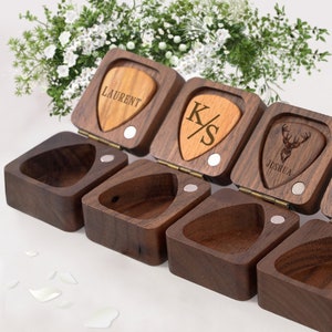 May include: A set of five wooden ring boxes with guitar pick shaped lids. The lids are engraved with names and initials. The boxes are made of dark wood and have a hinged lid.