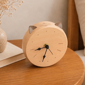 May include: A round light-coloured wooden analogue clock with two small cat-like ears on top. The clock face features black hands, simple hour markers, and the text "Happy Birthday" printed in a small font near the top edge of the wooden frame.