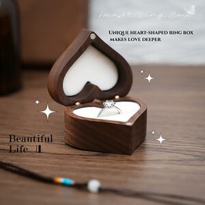 May include: A heart-shaped wooden ring box with a velvet interior. The box is open and a diamond ring is inside. The text "Unique heart-shaped ring box makes love deeper" is above the box. The text "Beautiful Life" is below the box.