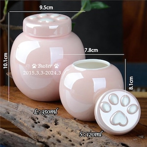 Pu&ograve; includere: Due urne per animali domestici in ceramica rosa con coperchi a forma di zampa. L'urna pi&ugrave; grande ha un diametro di 9,5 cm e un'altezza di 10,1 cm. L'urna pi&ugrave; piccola ha un diametro di 7,8 cm e un'altezza di 8,1 cm. L'urna pi&ugrave; grande ha il testo "Buster 2015.3.3-2024.3" sul davanti. Le urne sono entrambe decorate con impronte di zampe bianche.