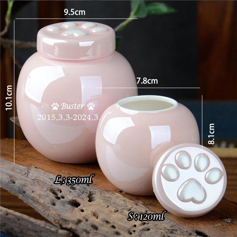 Pu&ograve; includere: Due urne per animali domestici in ceramica rosa con coperchi a forma di zampa. L'urna pi&ugrave; grande ha un diametro di 9,5 cm e un'altezza di 10,1 cm. L'urna pi&ugrave; piccola ha un diametro di 7,8 cm e un'altezza di 8,1 cm. L'urna pi&ugrave; grande ha il testo "Buster 2015.3.3-2024.3" sul davanti. Le urne sono entrambe decorate con impronte di zampe bianche.