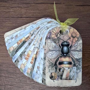 May include: A decorative tag with a bee illustration. The tag features a detailed drawing of a bee with a honeycomb background and floral accents. The tag is attached to a fan of patterned paper with a yellow ribbon.