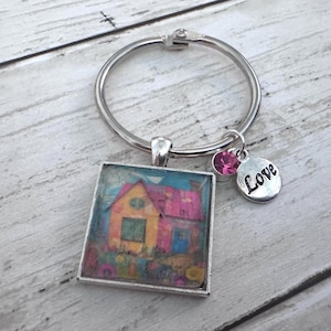 May include: A silver-toned metal keyring featuring a square pendant with a colorful illustration of a house with a pink roof and yellow walls. Attached to the ring is a small round charm with the word Love engraved and a single pink crystal bead.