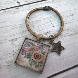 May include: A bronze-toned metal keyring featuring a circular ring, a square pendant, and a star-shaped charm. The square pendant displays a graphic of sunflowers, butterflies, and an American flag. The star charm also features a flag pattern.